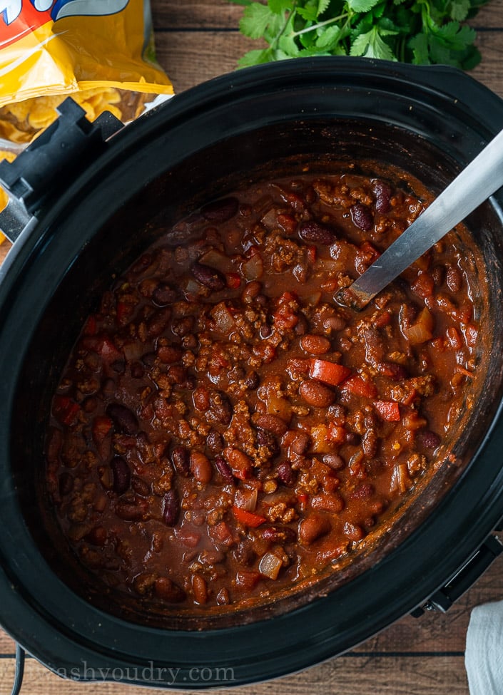 black slow cooker with chili inside with spoon and frito chips beside it.