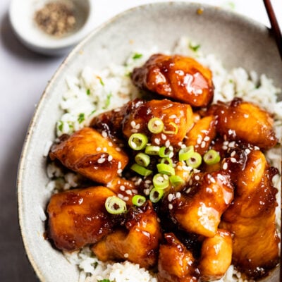 bowl of crispy sesame chicken on top of white rice with chopsticks.