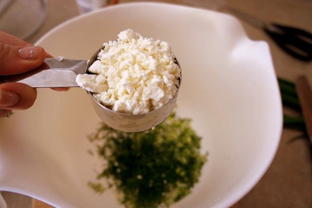 A measuring cup of feta cheese over a bowl of chopped dill and scallions