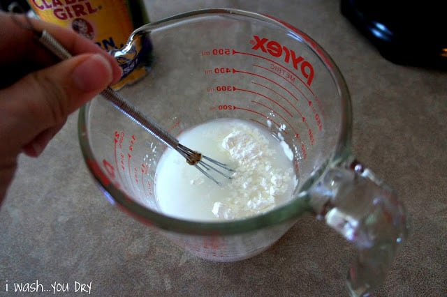 A hand using a small whisk to combine water and cornstarch in a glass measuring cup