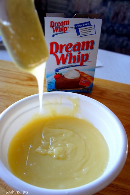 A spatula dripping glaze over a bowl of glaze, in front of a container of Dream Whip