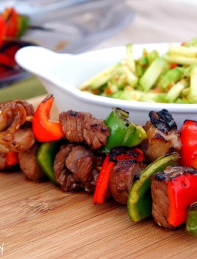 A close up of a two steak cubes and pepper slices on a skewers displayed in front of a bowl of veggies.