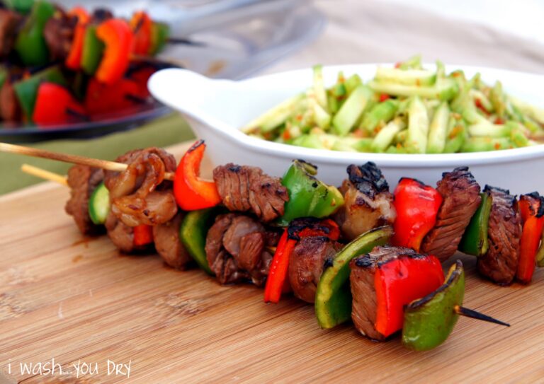 A close up of a two steak cubes and pepper slices on a skewers displayed in front of a bowl of veggies.  