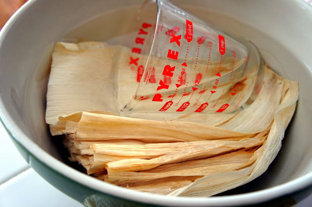 Corn husks in a pan of water with a glass measuring cup adding weight to hold them down