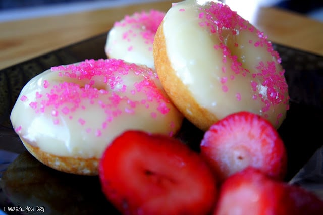 A close up of vanilla glazed donuts, with pink sprinkles, on a plate with a side of strawberries