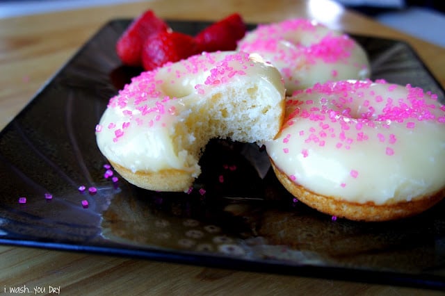 Frosted baked donuts displayed on a plate topped with pink sprinkles; one donut with a bite taken from it