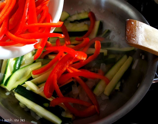Sliced peppers being added to a pan of sautéing veggies.
