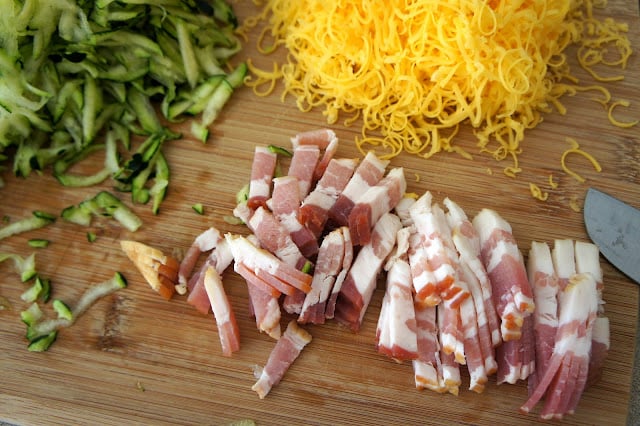 A pile of raw bacon sliced into thin strips on a cutting board next piles of shredded cheese and zucchini