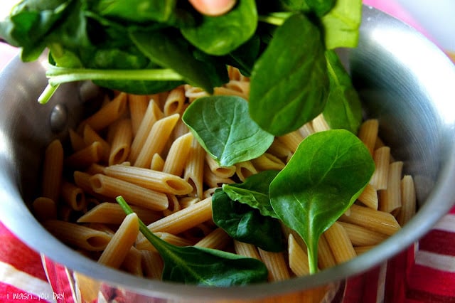 Spinach leaves being added to a pan of cooked penne pasta