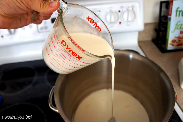 A hand pouring a measuring cup with milk and cream into a pot on the stove