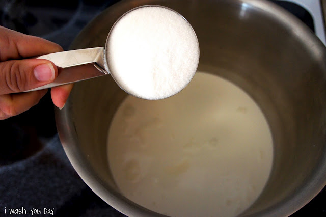 A hand pouring a measuring cup of sugar in to a pot of milk and cream