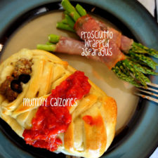 A mummy shaped calzone, topped with a little sauce on a plate next to bacon wrapped asparagus