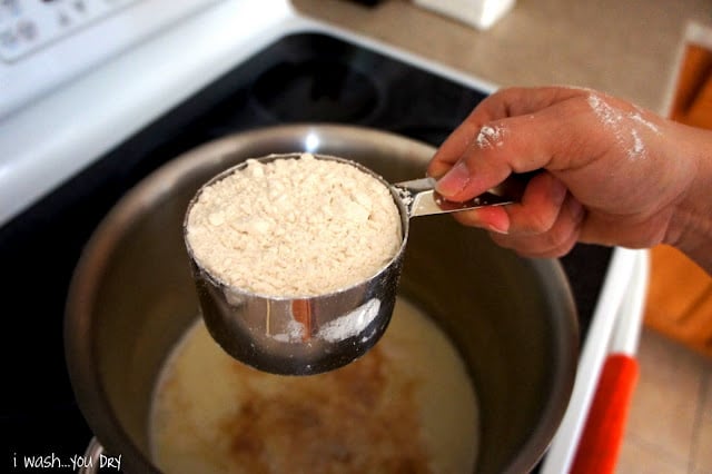 A hand adding a measuring cup of flour to the pot of liquid