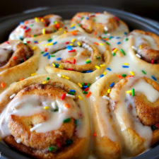 A close up of cinnamon rolls in a round pan topped with icing and sprinkles