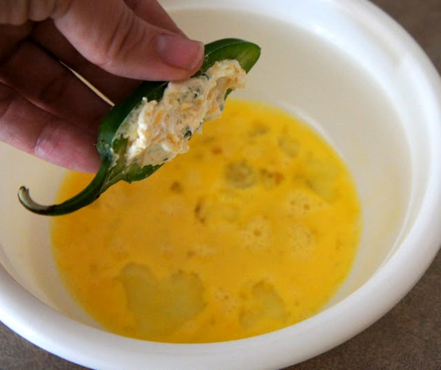 A hand dipping a cheese filled jalapeño half into a bowl of lightly beaten egg