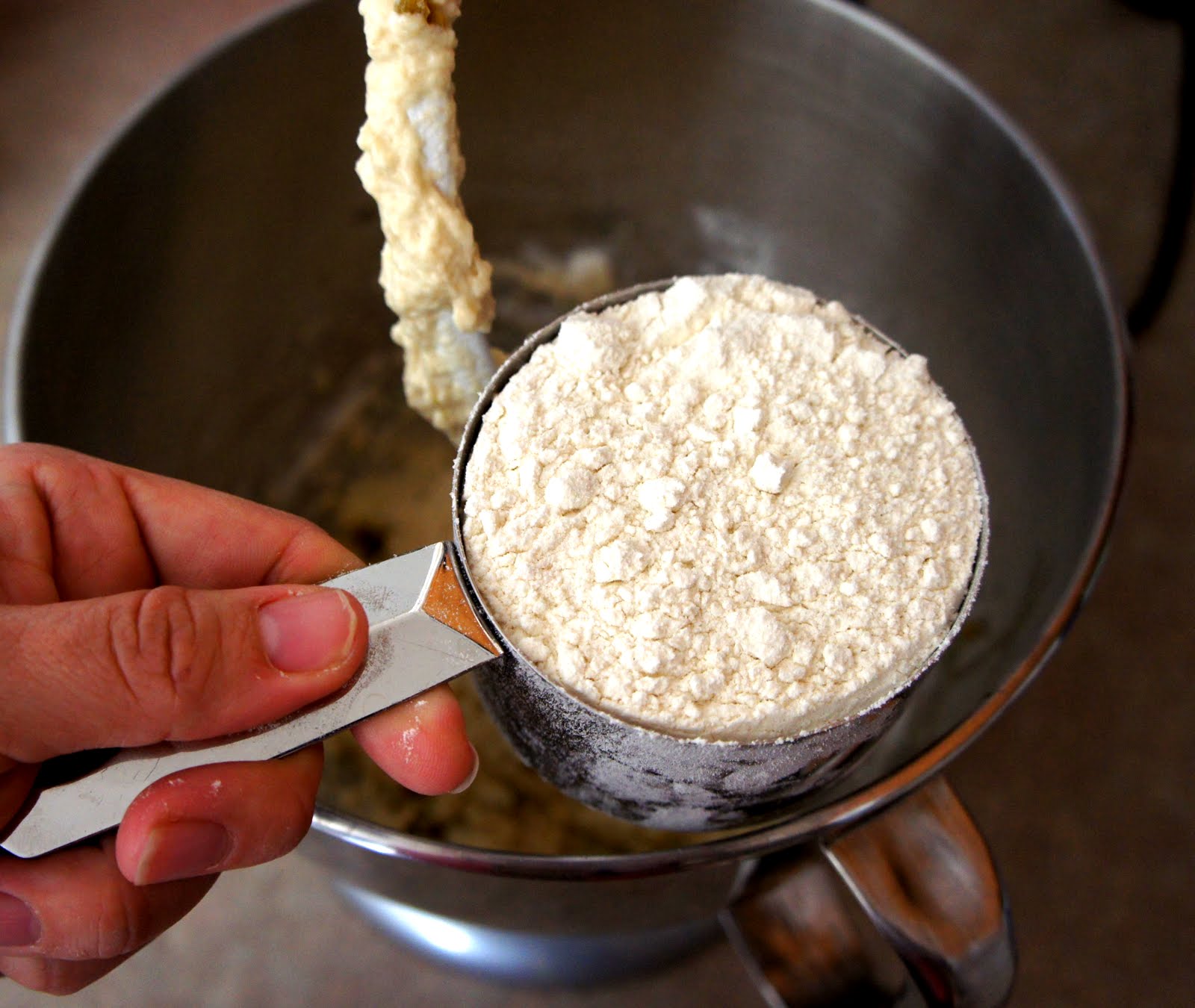 A hand holding a measuring cup of flour above a mixing bowl