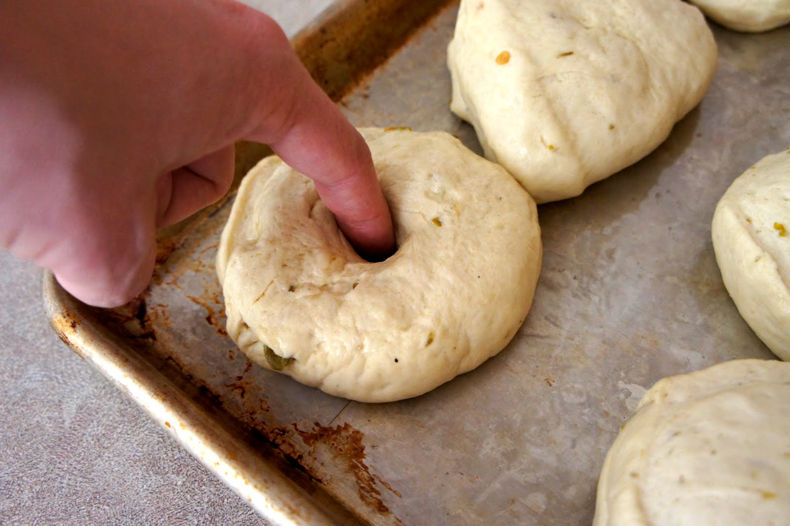 A close up of raw bagel dough on a pan, with a hand making a center hole