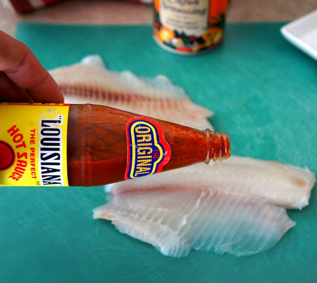 A hand holding a bottle of Louisiana Hot Sauce in front of a couple pieces of raw Tilapia on a cutting board