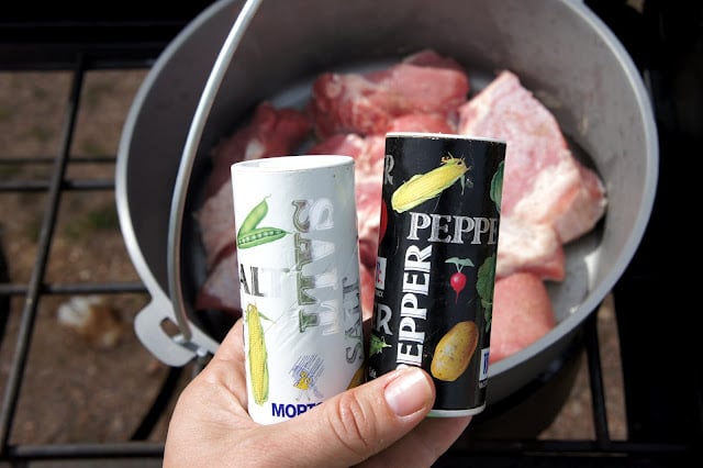 A hand holding bottles of salt and pepper in front of a Dutch Oven filled with raw rib meat
