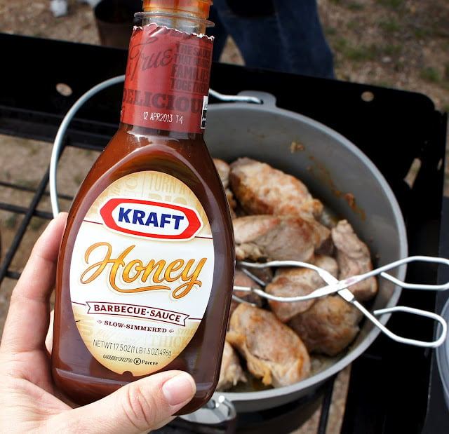 A hand holding a bottle of Kraft\'s Honey Barbecue Sauce in front of a Dutch Oven of cooked ribs