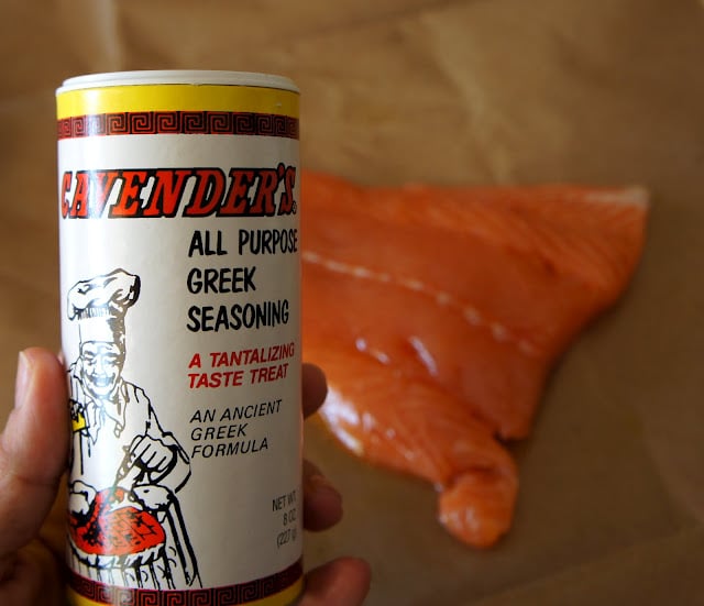 A close up of a hand holding a bottle of Greek seasoning in front of a salmon fillet