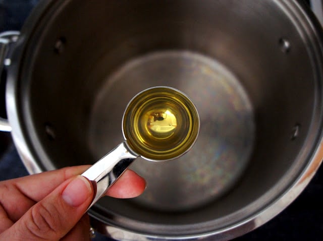A hand holding a measuring spoon of oil over a pot