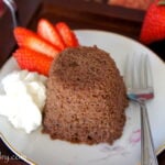 A close up of a chocolate mug cake displayed on a plate next to a sliced strawberry and some whipped topping