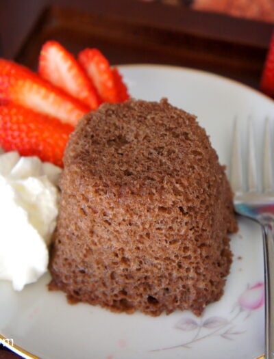A close up of a chocolate mug cake displayed on a plate next to a sliced strawberry and some whipped topping