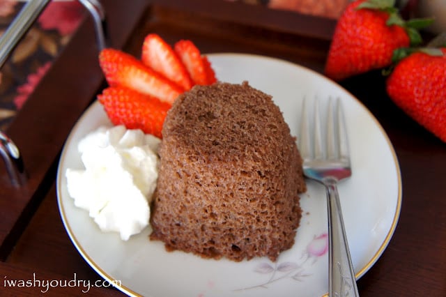 A close up of a chocolate mug cake displayed on a plate next to a sliced strawberry and some whipped topping