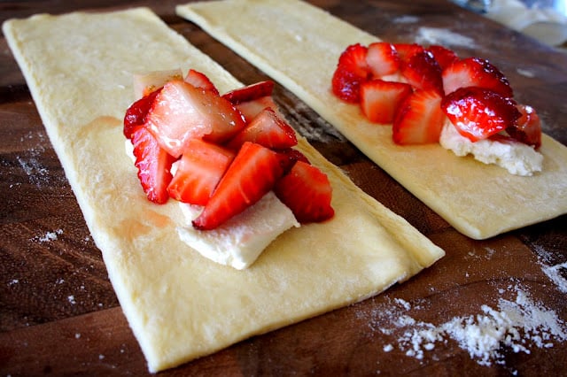 Two strips of pastry dough on a cutting board, each topped with some cream cheese and sliced strawberries