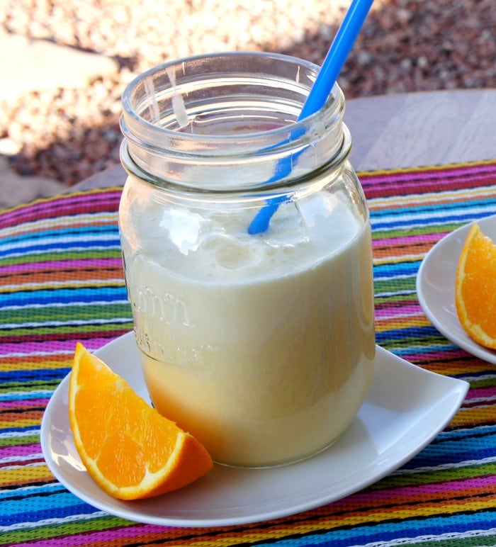 A close up of Pineapple Orange Julius in a mason jar glass on a table next to a slice of orange
