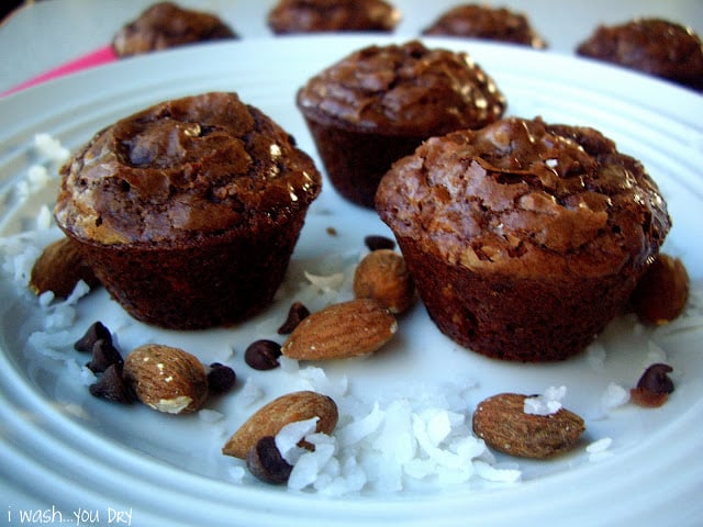 Three brownie muffins on a plate surrounded by chocolate chips, almonds and coconut flakes. 