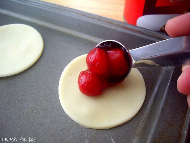 A spoon adding cherry pie filling on top of a mini circle of pie crust on a pan.