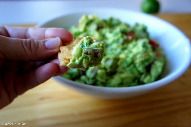 A hand holding a chip with guacamole on it in front of a bowl of guacamole.