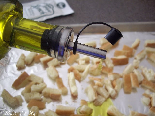 Olive oil being drizzled onto a baking sheet with small bread pieces. 