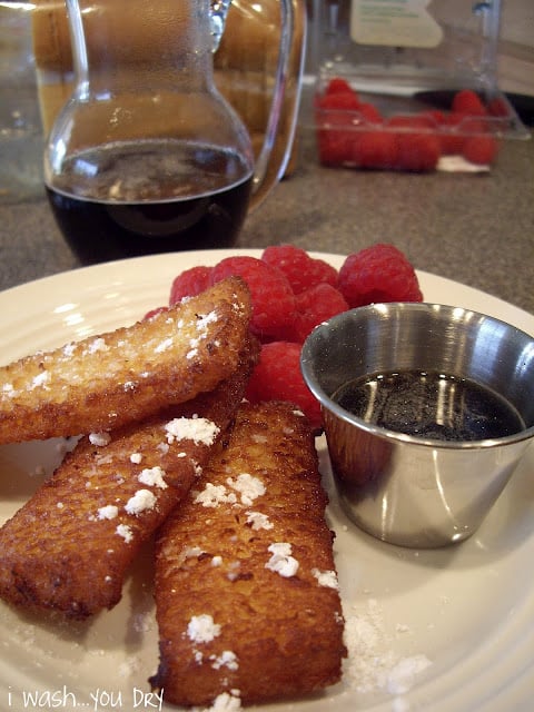 A display of french toast strips sprinkled with powdered sugar with a side of raspberries and syrup. 