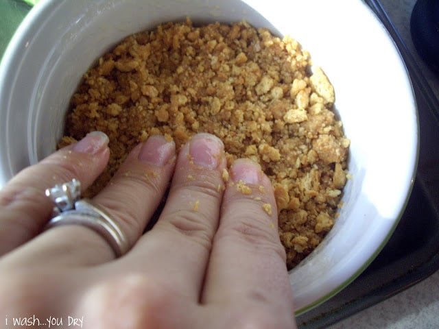 A hand pressing the graham cracker mixture into the bottom of a small glass dish. 