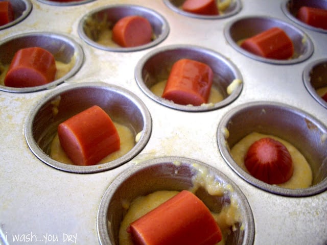 A muffin tin with a hotdog chunk added to the top of each spot on top of some muffin batter. 