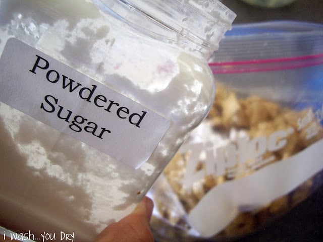 A hand holding a container of powdered sugar in front of a ziplock bag with cereal mixture in it. 