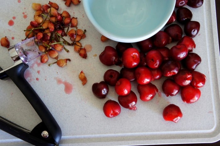 A close up of a cutting board with a pile of cherry seeds, a pile of cherries and an empty bowl