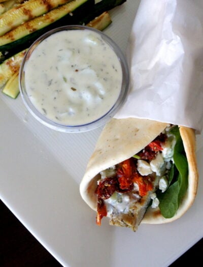 A wrapped Grilled Chicken Souvlaki Pita on a plate next to a small bowl of dipping sauce and grilled zucchini strips