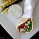 A wrapped Grilled Chicken Souvlaki Pita on a plate next to a small bowl of dipping sauce and grilled zucchini strips