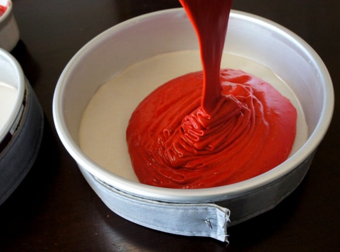 A close up of a round cake pan, with red cake batter being poured on top of white cake batter