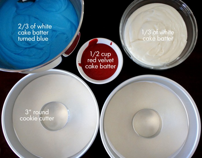 A close up of 3 different bowls of colored cake batter (blue, red and white) and two empty round cake pans