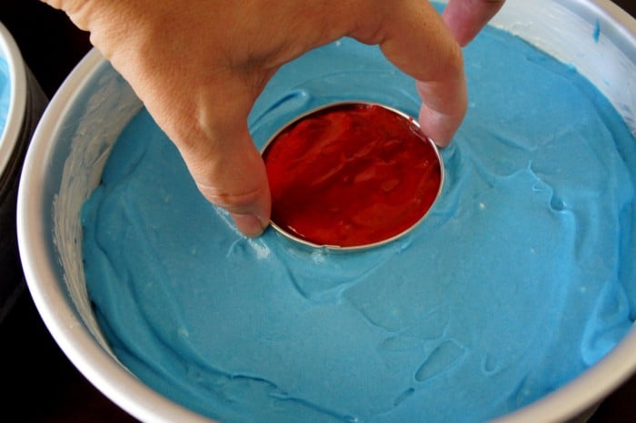 A close up of a round cake pan with blue cake batter and a hand inserting a small center pan with red cake batter