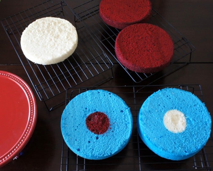 5 cake layers cooling on a cooling rack, 2 red layers, 2 blue layers with different colored centers and one white layer