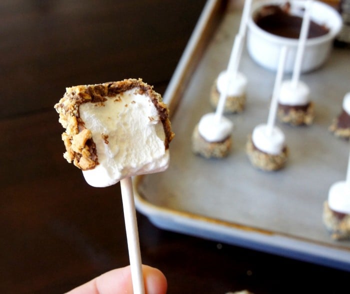 A S\'more\'s marshmallow pop with a a bite taken from it in front of a pan of already made S\'mores pops