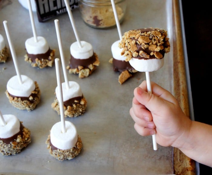 A small hand holding a S\'more dipped marshmallow pop in front of a pan of marshmallow pops
