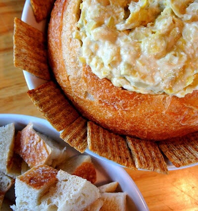 A close up of a display of a bread bowl with a dip inside of it, lined with crackers and next to a bowl of chopped bread.