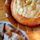A close up of a display of a bread bowl with a dip inside of it, lined with crackers and next to a bowl of chopped bread.